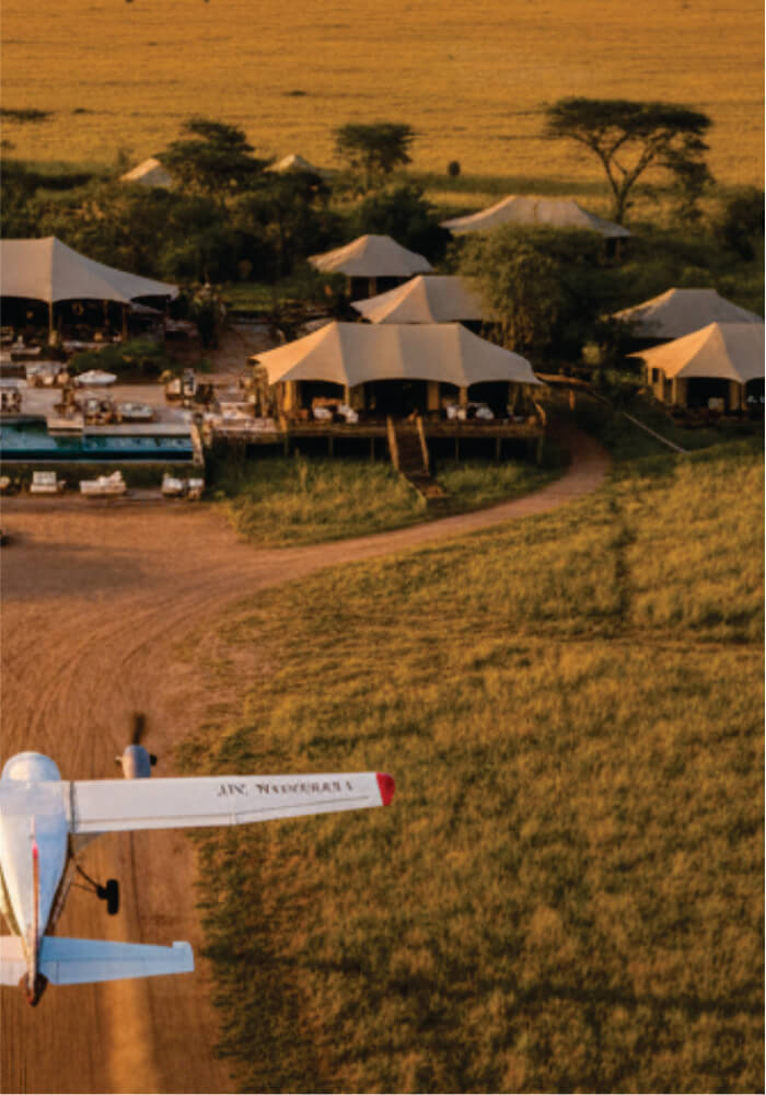 A scenic view of a safari camp with tents and an airplane in the foreground.