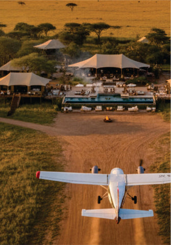 Small airplane on a dirt airstrip with safari tents and vehicles in the background.
